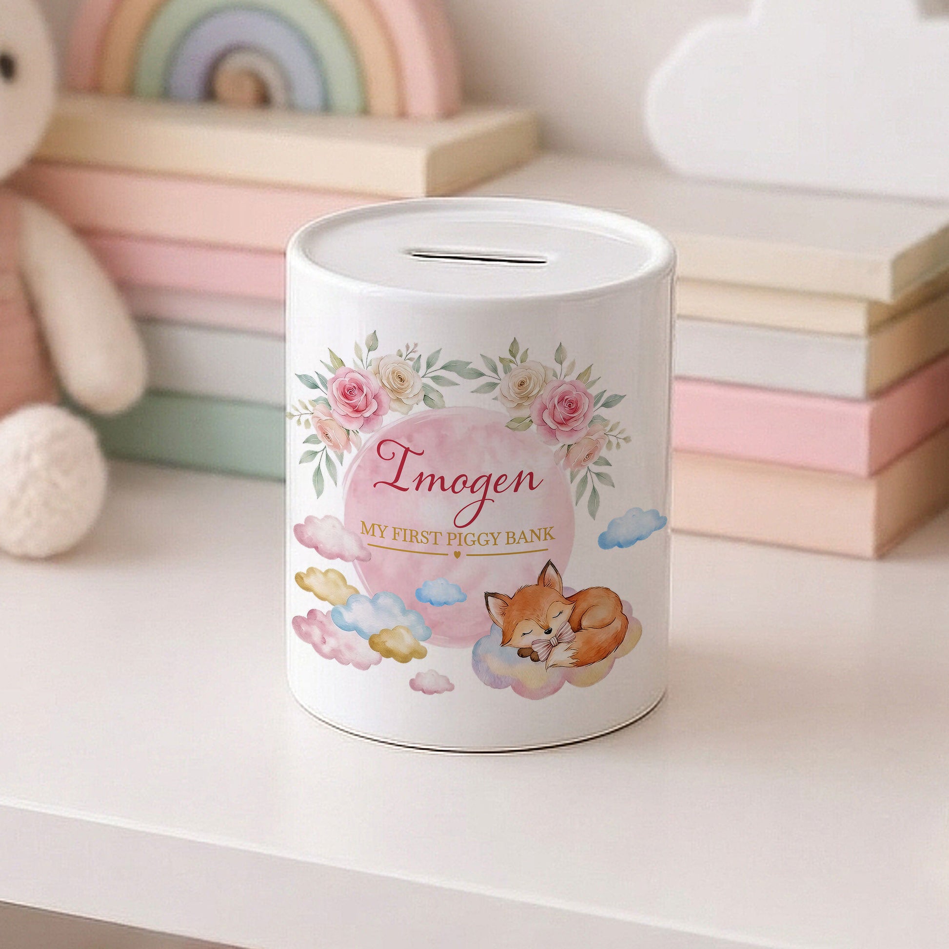 A white cylindrical piggy bank with a slot on top, decorated with pink flowers, a sleeping fox, clouds, and the text 'Imogen MY FIRST PIGGY BANK'. It sits on a white surface with pastel-coloured books and a plush toy blurred in the background.