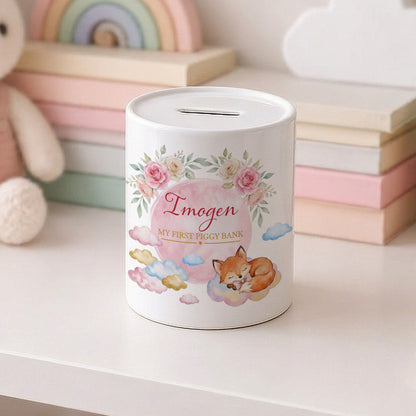 A white cylindrical piggy bank with a slot on top, decorated with pink flowers, a sleeping fox, clouds, and the text 'Imogen MY FIRST PIGGY BANK'. It sits on a white surface with pastel-coloured books and a plush toy blurred in the background.
