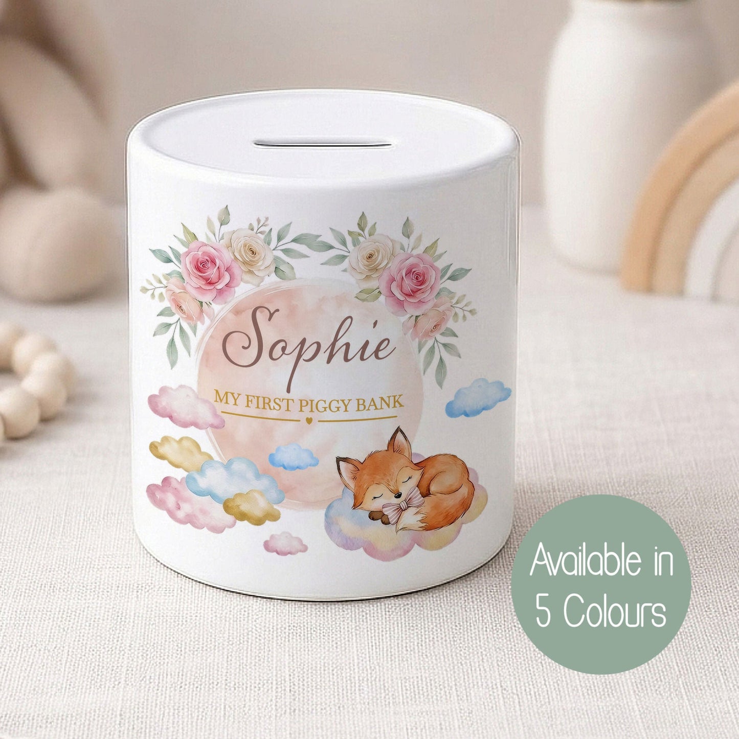 A personalised white piggy bank with a floral and fox illustration, labelled 'Sophie MY FIRST PIGGY BANK', shown on a soft neutral fabric surface with blurred nursery decor in the background. A green circle badge reads 'Available in 5 Colours'.