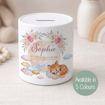 A personalised white piggy bank with a floral and fox illustration, labelled 'Sophie MY FIRST PIGGY BANK', shown on a soft neutral fabric surface with blurred nursery decor in the background. A green circle badge reads 'Available in 5 Colours'.