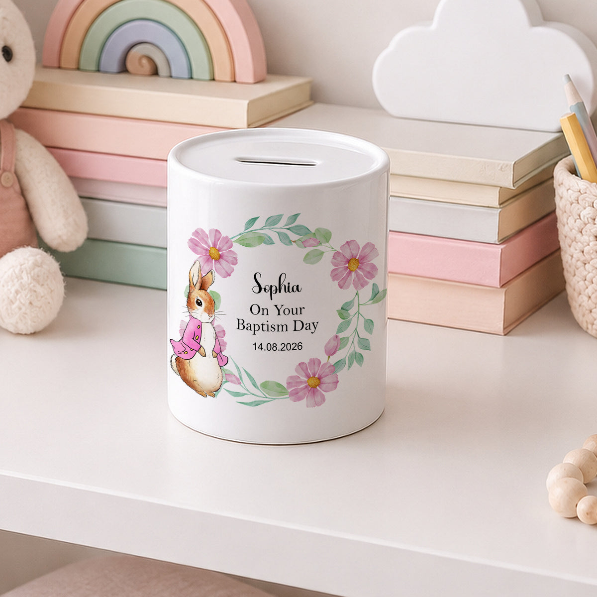 A white cylindrical baptism money box with a slot on top, decorated with a pink bunny, pink flowers, green leaves, and personalized text reading 'Sophia On Your Baptism Day 14.08.