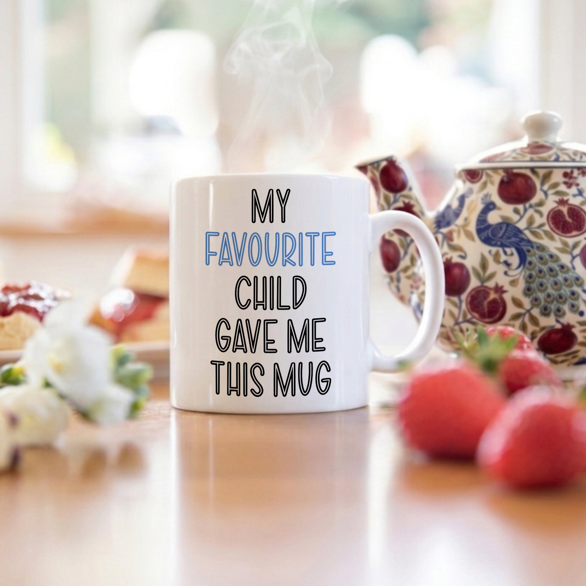 White mug with text 'My favourite child gave me this mug' on a table with a teapot and fruit in the background.
