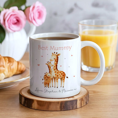 A white ceramic mug with a custom 'Best Mummy' giraffe illustration sits on a wooden coaster on a wooden table, surrounded by breakfast items like croissants and orange juice.