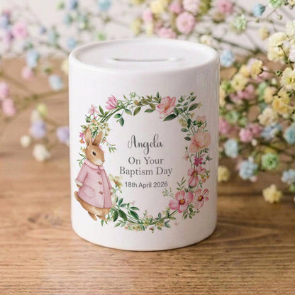 A white ceramic baby baptism money box with a personalised illustration of a bunny in a floral wreath, sitting on a wooden surface with baby's breath flowers in the background.