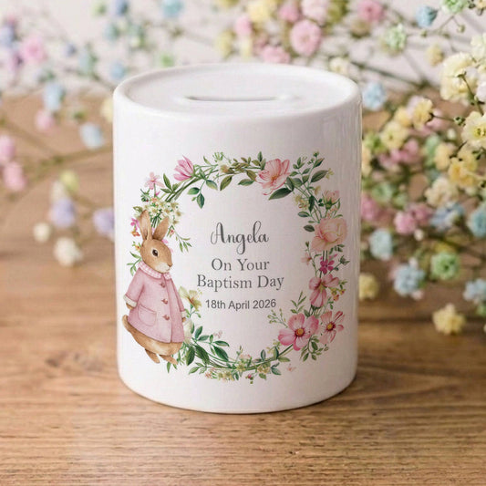 A white ceramic baby baptism money box with a personalised illustration of a bunny in a floral wreath, sitting on a wooden surface with baby's breath flowers in the background.