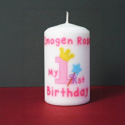 A white birthday candle with pink text reading 'Imogen Rose My 1st Birthday' and a decorative pink number 1 with a yellow crown, blue star, and wand, shown on a red surface against a dark grey background.