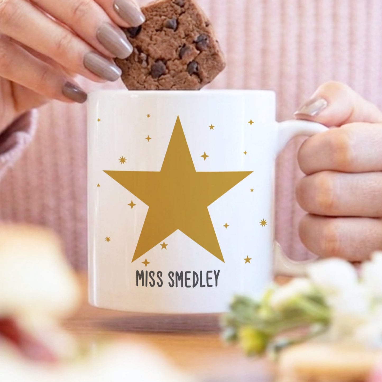 Person holding a mug with a gold star design and 'MISS SMEDLEY' text, eating a cookie.