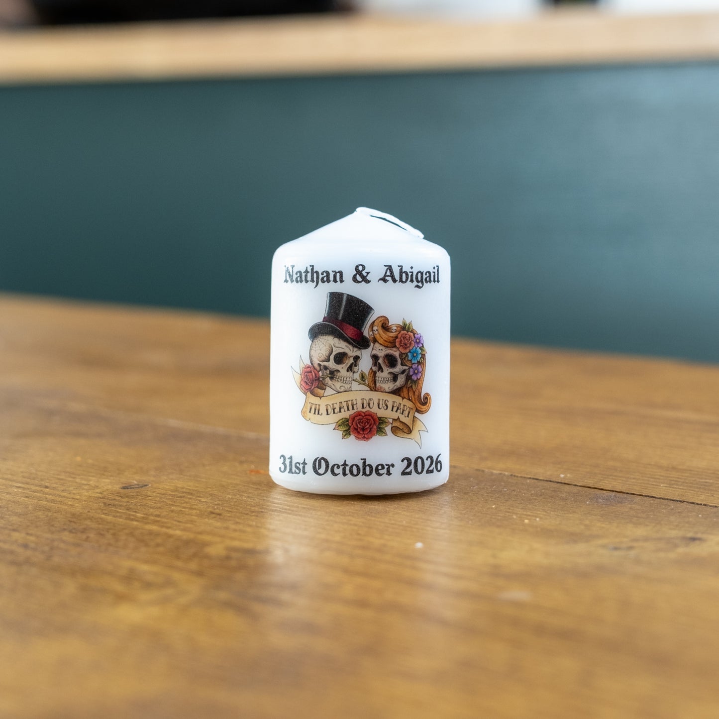 A small white personalised candle standing on a wooden tabletop, printed with the names Nathan & Abigail, a skull illustration with a top hat and roses, and the date 31st October 2026.