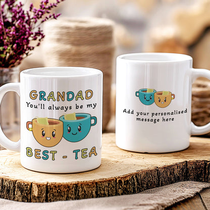 Two white ceramic mugs displayed on a rustic wood slice. The mug on the left is personalised with the text 'GRANDAD You'll always be my BEST - TEA' and features two smiling cartoon cups.