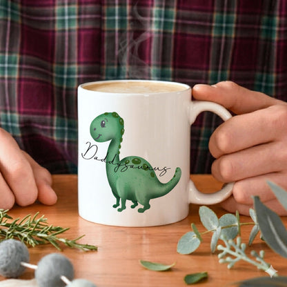 Person holding a mug with a dinosaur design and 'Dad'saurus' text on a wooden table.