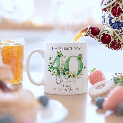 Personalized birthday mug with floral design and text, surrounded by a blurred background of a table with food and drinks.