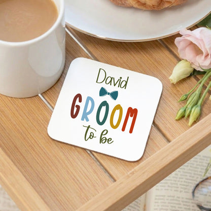 A square white coaster with rounded corners on a wooden table, printed with the text 'David GROOM to be' and a small bow tie graphic. A white mug, a plate with a pastry, and pink flowers are arranged around it in a styled setting.