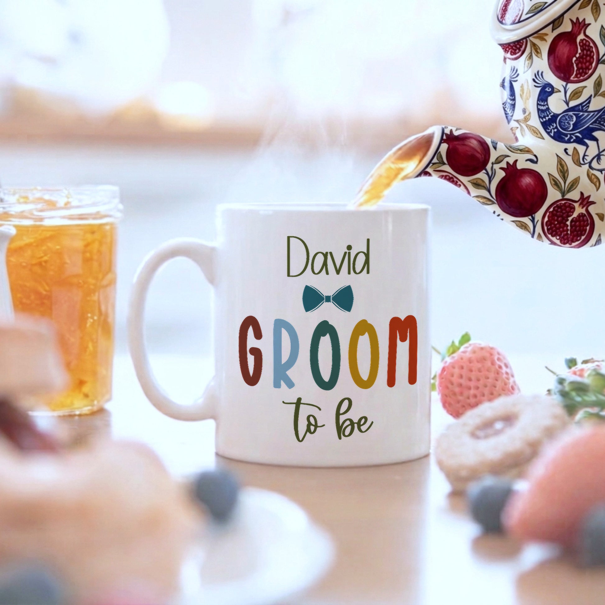 A white ceramic mug with the text 'David GROOM to be' and a small bow tie graphic, shown on a table with a teapot pouring tea, a jar of fruit, and pastries in a breakfast setting.