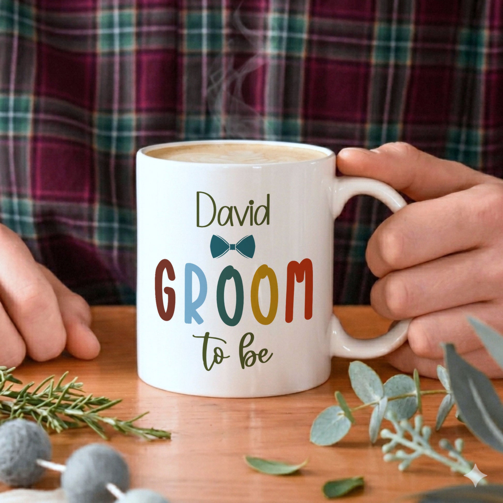 A white ceramic mug with the personalised text 'David Groom to be' and a small bow tie graphic, held by two hands over a wooden table with a plaid shirt in the background.