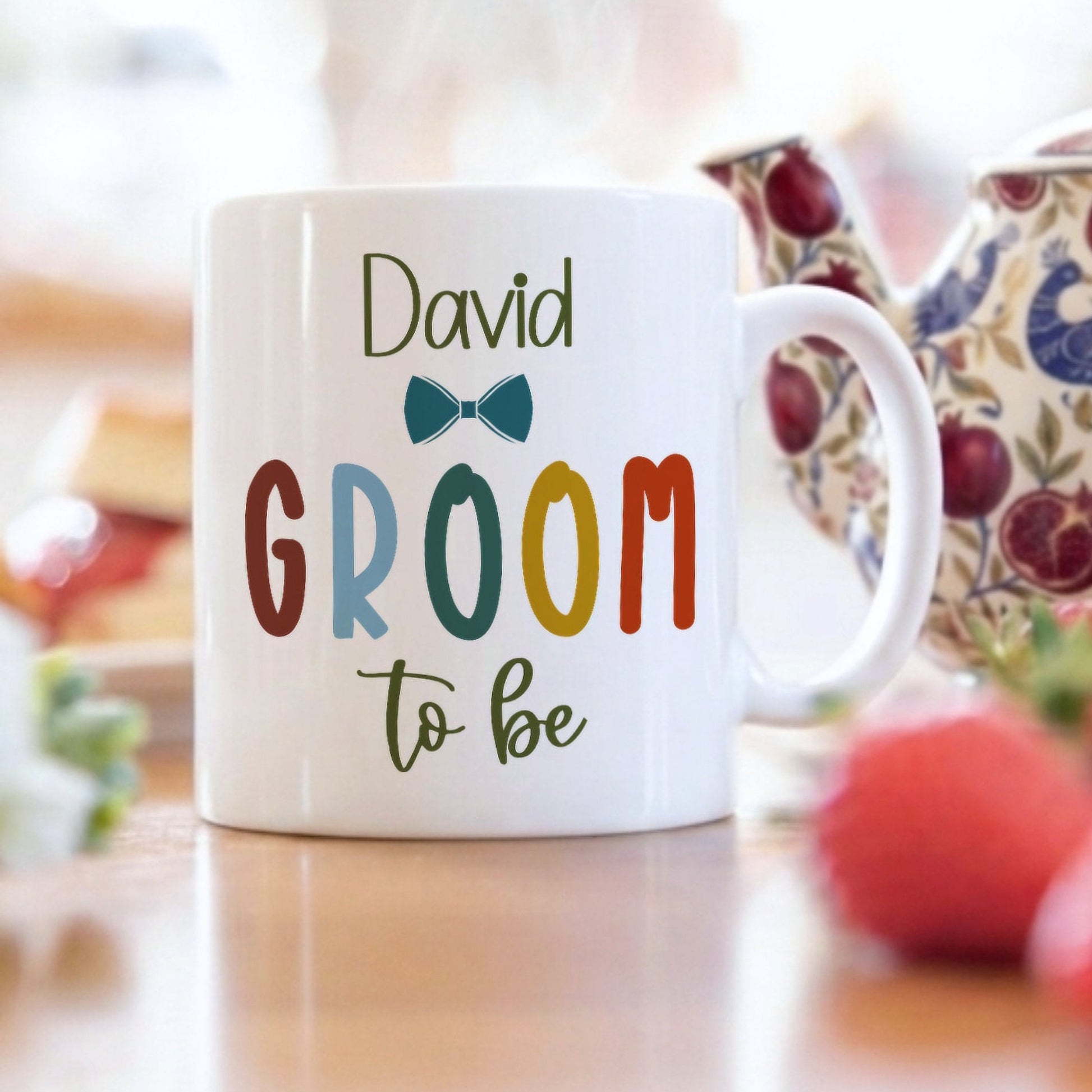 A white ceramic mug with the text 'David GROOM to be' printed on it, styled on a wooden table with a teapot, strawberries, and pastries in a soft-focus background.