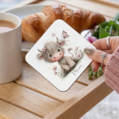 A square white coaster with a cartoon Highland cow design, adorned with a floral crown and pink flowers. The word "Mum" is written in cursive. The coaster is held near a tray with a croissant and a cup of tea.