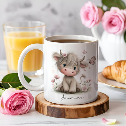 White ceramic mug with a cartoon Highland cow design, adorned with a pink flower crown and butterflies. The mug has the name "Jasmine" written on it. The mug sits on a wooden coaster, with a glass of orange juice, a rose, and a croissant in the background.