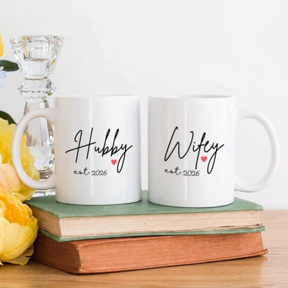 Two white mugs with 'Hubby' and 'Wifey' text on a wooden surface with flowers and books.