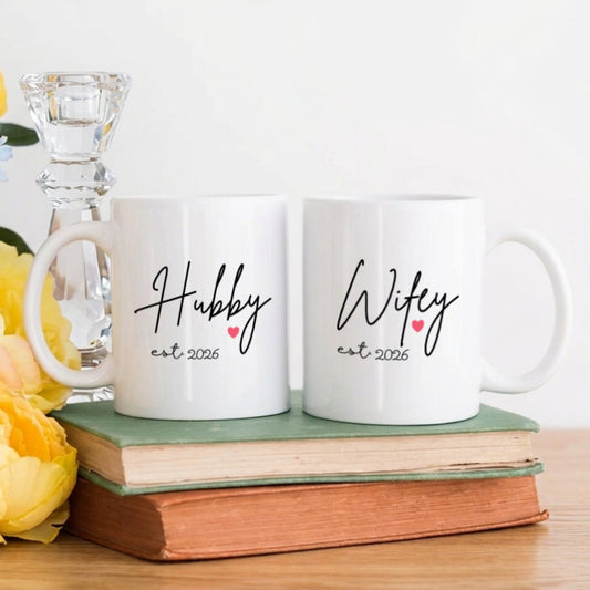 Two white mugs with 'Hubby' and 'Wifey' text on a wooden surface with flowers and books.
