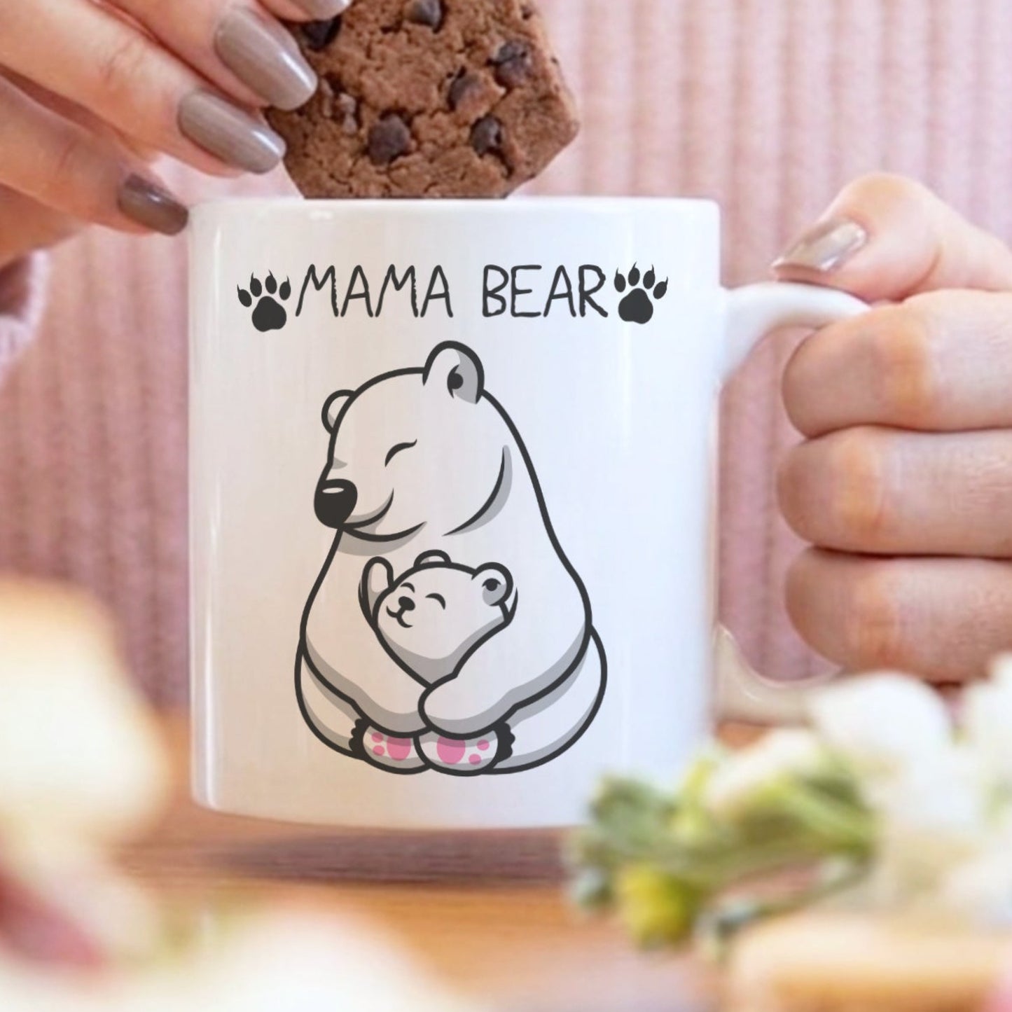 A white ceramic mug with a black line art illustration of a mama bear and cub, with the text 'MAMA BEAR' and paw prints. A person in a pink ribbed sweater is holding the mug while placing a chocolate chip cookie on top.