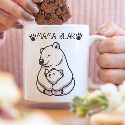 A white ceramic mug with a black line art illustration of a mama bear and cub, with the text 'MAMA BEAR' and paw prints. A person in a pink ribbed sweater is holding the mug while placing a chocolate chip cookie on top.