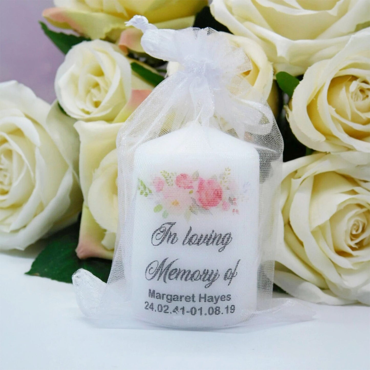 A white memorial candle in a clear tulle gift bag, decorated with pink flowers and the text 'In loving Memory of Margaret Hayes' and a date, set against a background of pale yellow roses.