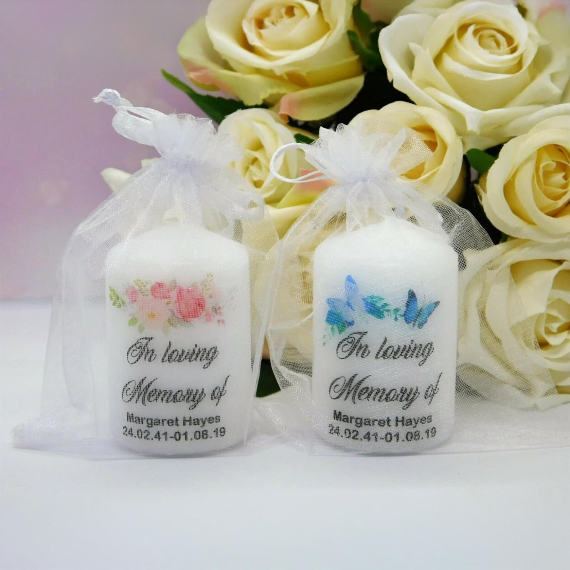 Two white memorial taper candles in clear organza pouches, decorated with floral and butterfly designs and printed with the text 'In loving Memory of Margaret Hayes' and a date, displayed in front of yellow roses.