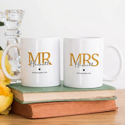 Two white ceramic mugs printed with 'MR & MRS Heather 1975 September 18th' in gold and black lettering, displayed on a stack of books next to yellow flowers and a glass candlestick.