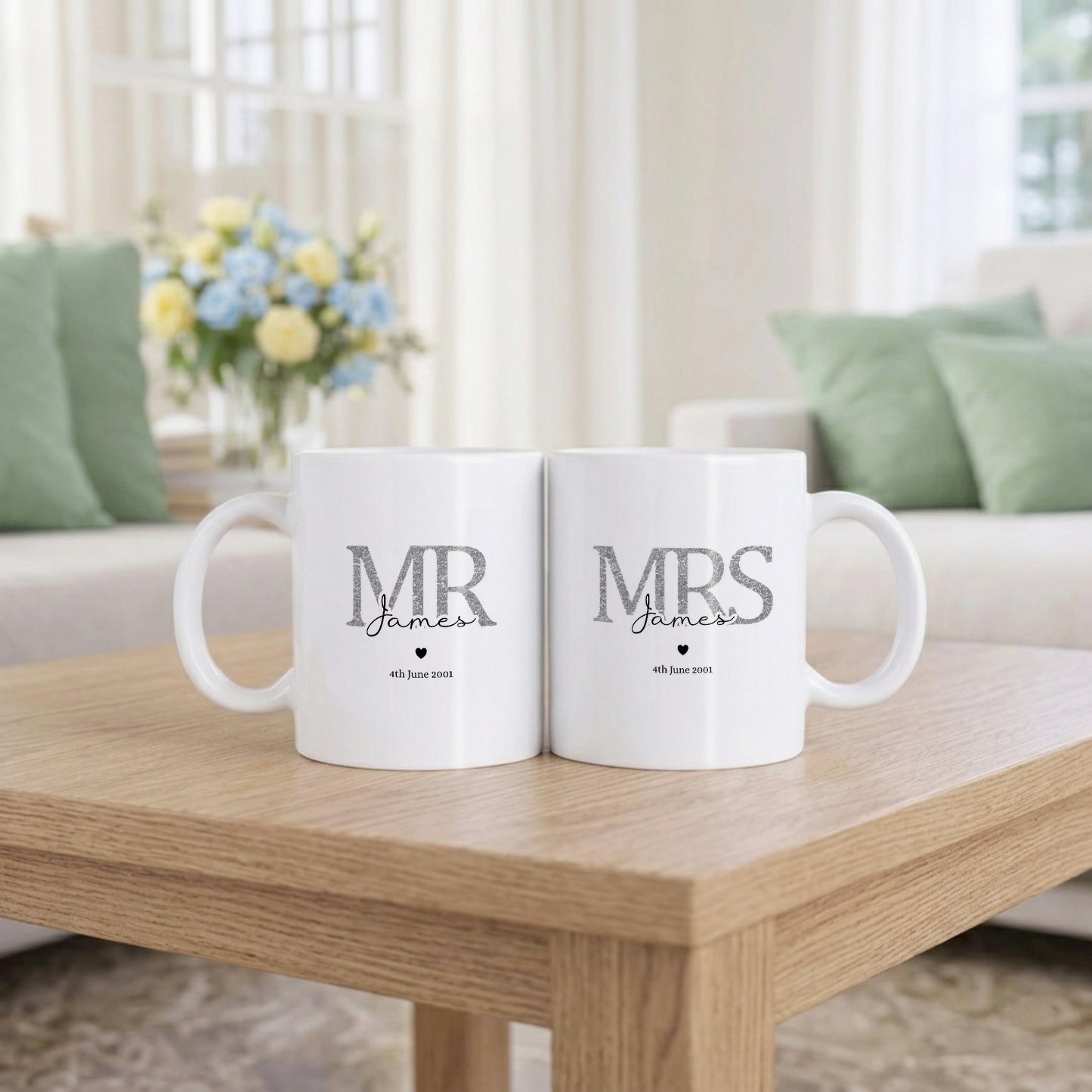 Two white ceramic mugs sitting side by side on a wooden table in a bright living room. The left mug is printed with 'MR James' and the right mug is printed with 'MRS James' in gray text with a small heart and date underneath.