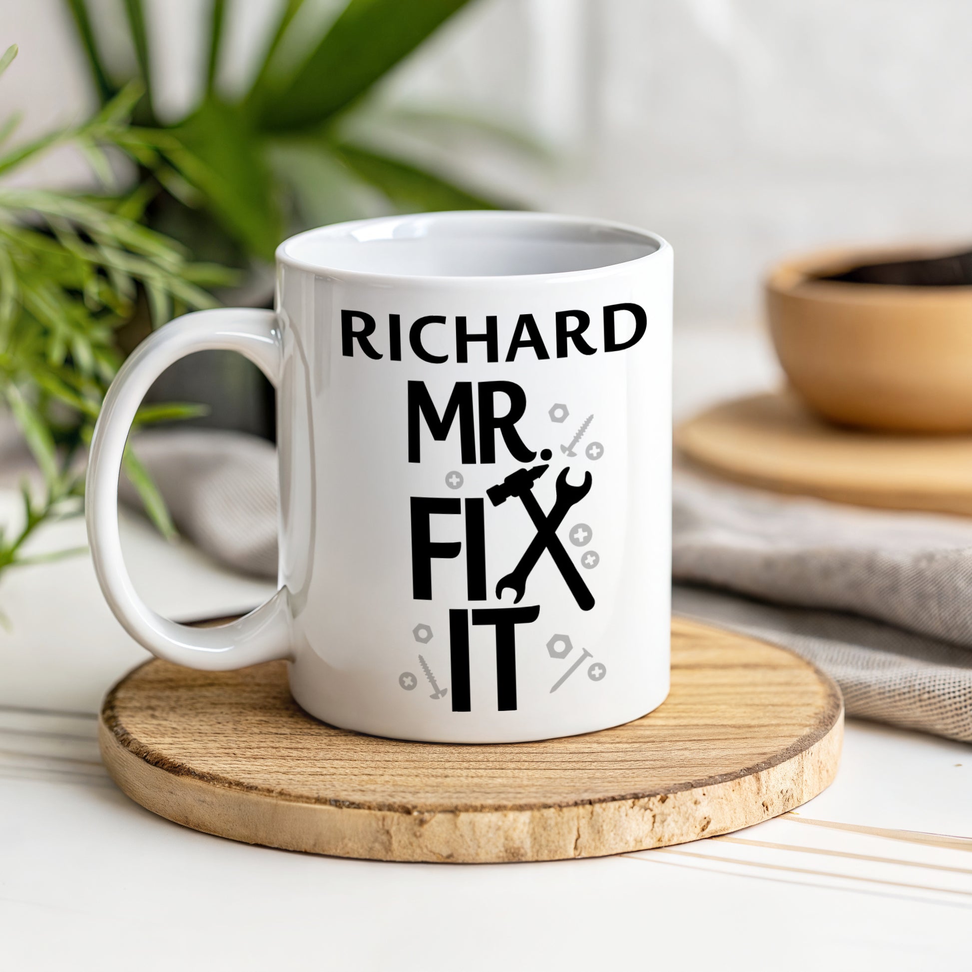 White mug with 'Richard Mr Fix It' text on a wooden coaster with plants and bowls in the background