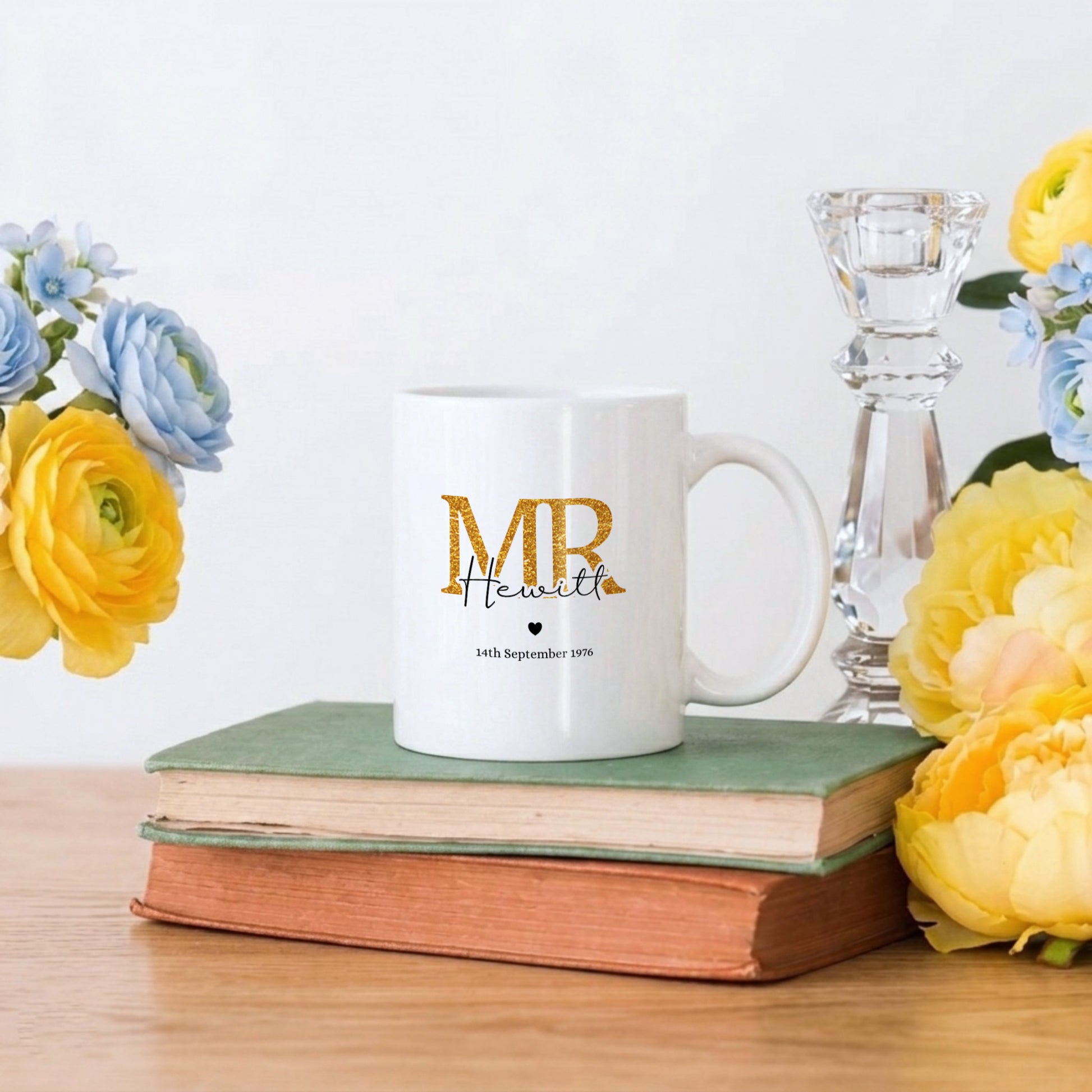 A white ceramic mug with gold and black personalized text sits on a stack of two vintage books on a wooden table, styled with yellow and blue flowers and a clear glass candlestick holder against a white background.