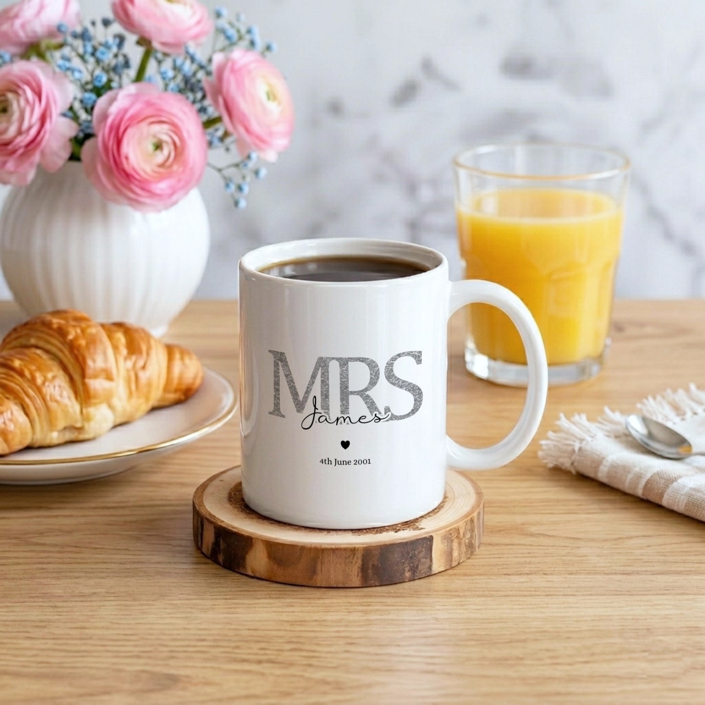 A white ceramic coffee mug with 'MRS James' printed on it sits on a wooden coaster on a wooden table, surrounded by a croissant, a glass of orange juice, and pink flowers in the background.