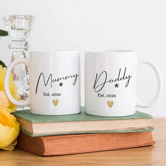 Two white mugs with 'Mummy Est. 2026' and 'Daddy Est. 2026' text on a wooden surface with books and flowers.