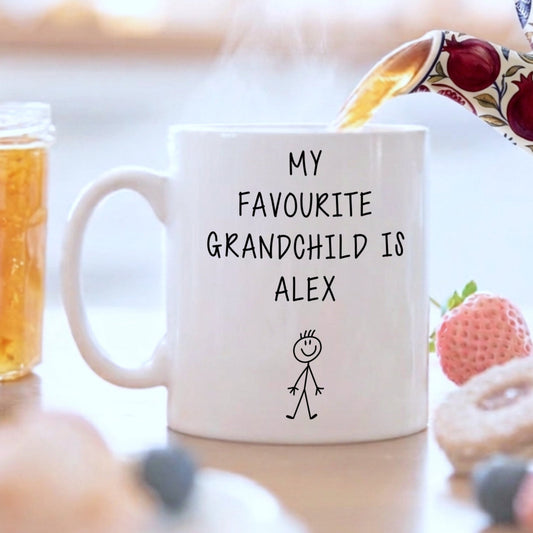 White mug with 'My favourite grandchild is Alex' text on a blurred background of food and drinks.