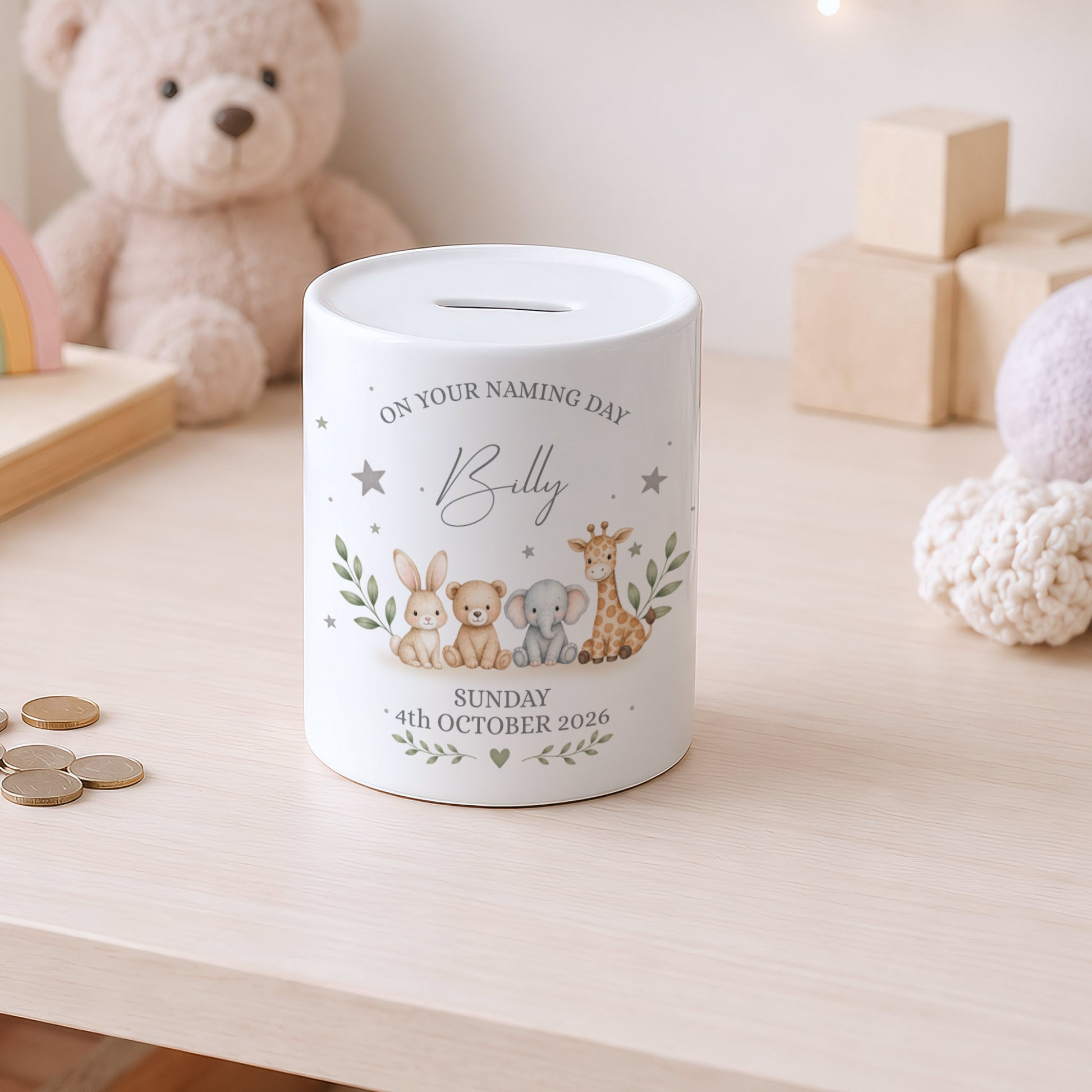 A white ceramic baby christening money box with a custom printed cartoon animal design and text, sitting on a light wooden table with a teddy bear in the background.