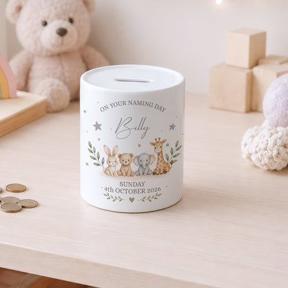 A white ceramic baby christening money box with a custom printed cartoon animal design and text, sitting on a light wooden table with a teddy bear in the background.