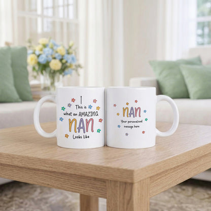 Two white ceramic mugs showing front and back of the Amazing Nan personalised mug design with colourful daisy flowers, displayed on a wooden coffee table in a bright living room