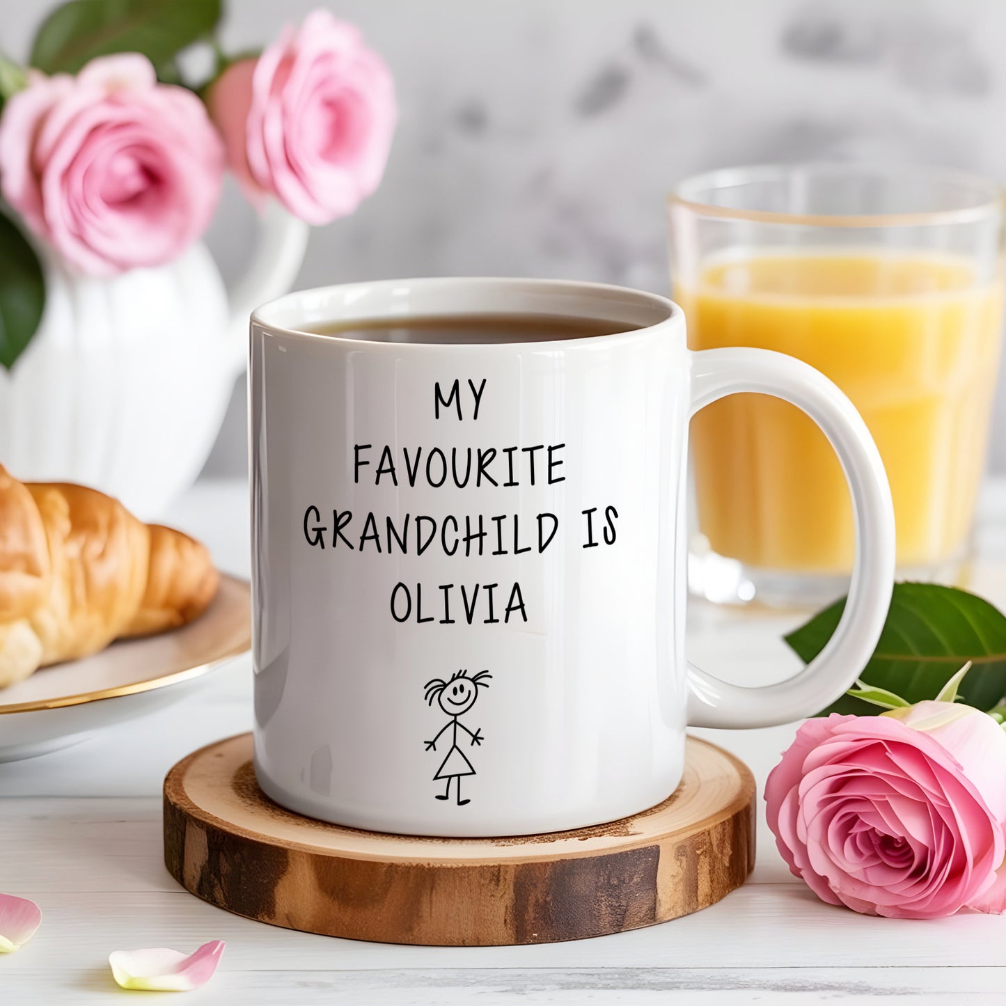 White mug with 'My favourite grandchild is Olivia' text on a wooden coaster with coffee, croissant, and flowers.