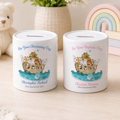 Two personalised ceramic piggy banks on a light wood surface, with a teddy bear, flowers, and pastel rainbow toy in the background. One bank is labelled 'On Your Christening Day' and the other 'On Your Baptism Day,' both with custom names and dates below a Noah's Ark illustration.