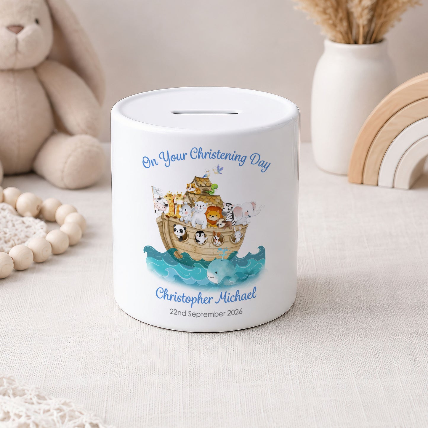 A white ceramic baby savings jar with a personalised christening illustration of Noah's Ark and the text 'On Your Christening Day Christopher Michael 22nd September 2026'. The jar is styled on a light beige fabric surface with a wooden bead necklace, a plush bunny, and a wooden rainbow in the background.