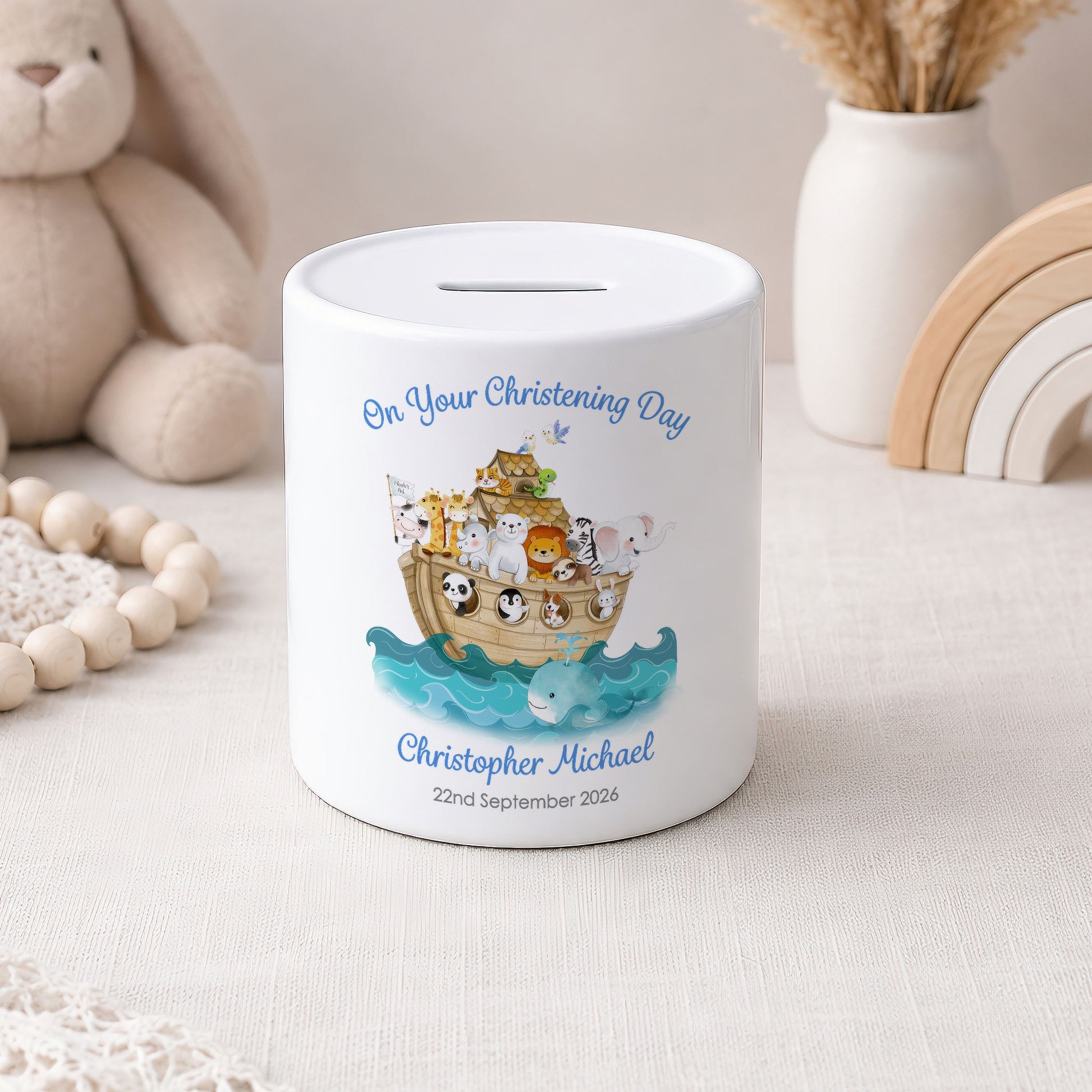 A white ceramic baby savings jar with a personalised christening illustration of Noah's Ark and the text 'On Your Christening Day Christopher Michael 22nd September 2026'. The jar is styled on a light beige fabric surface with a wooden bead necklace, a plush bunny, and a wooden rainbow in the background.