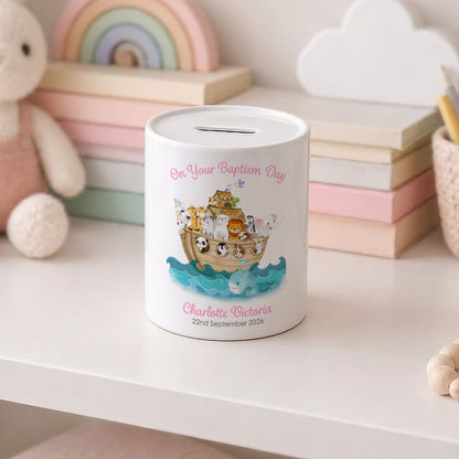 A white personalized baby baptism money box with a Noah's Ark illustration and the text 'On Your Baptism Day' and 'Charlotte Victoria 2nd September 2026', displayed on a white shelf in a nursery setting.