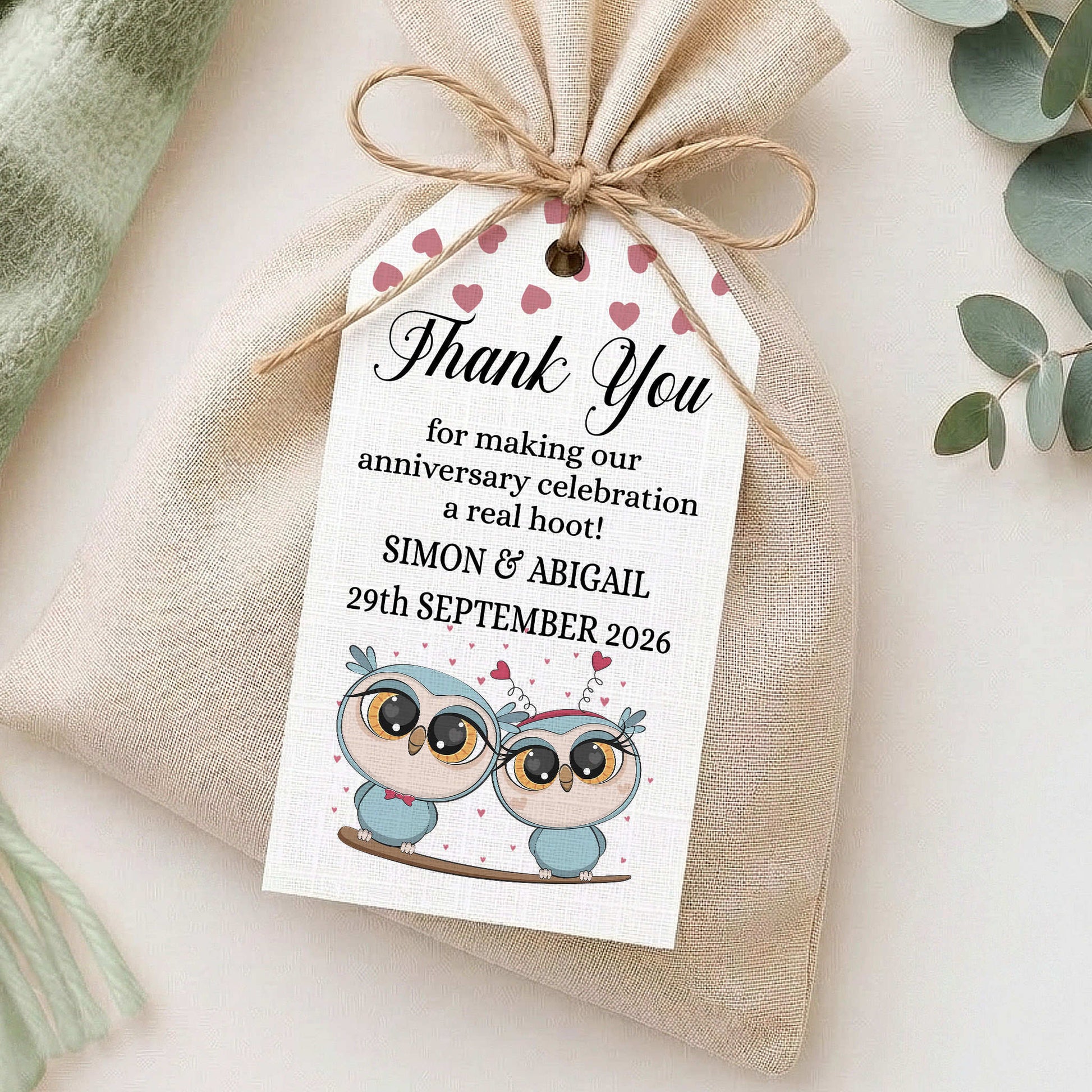 A small beige fabric gift bag tied with twine, featuring a white thank-you tag printed with pink hearts, owl illustrations, and personalised anniversary text, styled on a light surface with soft decorative props.