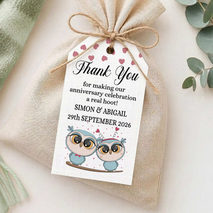 A small beige fabric gift bag tied with twine, featuring a white thank-you tag printed with pink hearts, owl illustrations, and personalised anniversary text, styled on a light surface with soft decorative props.