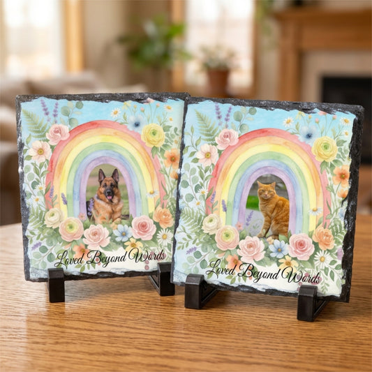 Two square slate coasters displayed on small black stands on a wooden table. Each coaster features a colorful rainbow, floral border, and a pet portrait: a German Shepherd on the left and an orange cat on the right, with the phrase 'Loved Beyond Words' printed at the bottom.