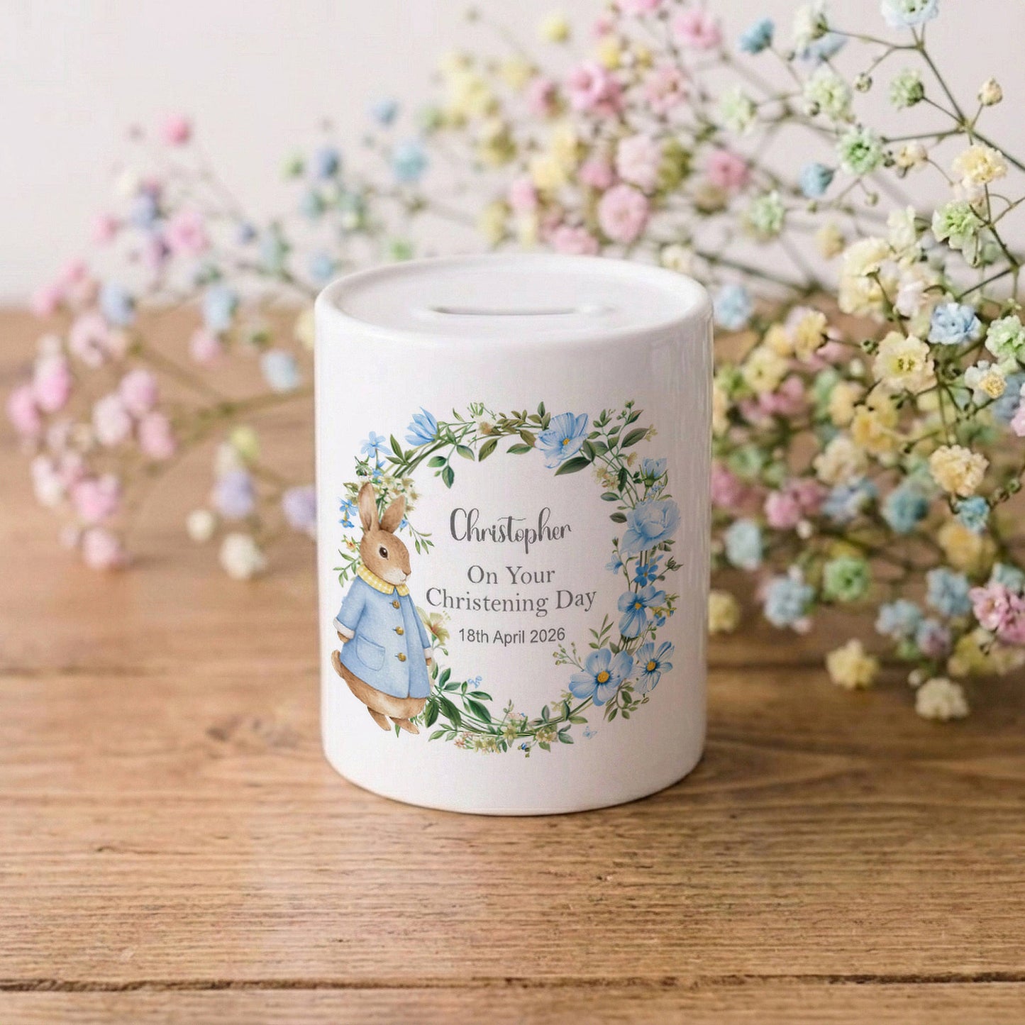 A white ceramic baby christening money box featuring a personalised illustration of Beatrix Potter's Peter Rabbit in a blue jacket, surrounded by a blue and green floral wreath. The box is printed with the name 'Christopher', 'On Your Christening Day', and a date.