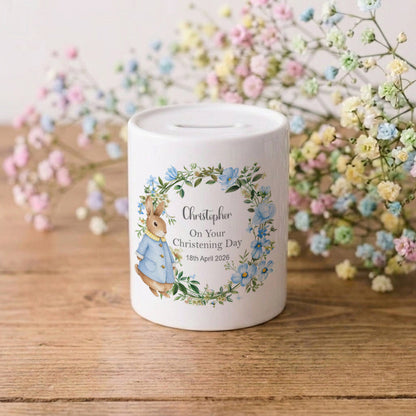 A white ceramic baby christening money box featuring a personalised illustration of Beatrix Potter's Peter Rabbit in a blue jacket, surrounded by a blue and green floral wreath. The box is printed with the name 'Christopher', 'On Your Christening Day', and a date.