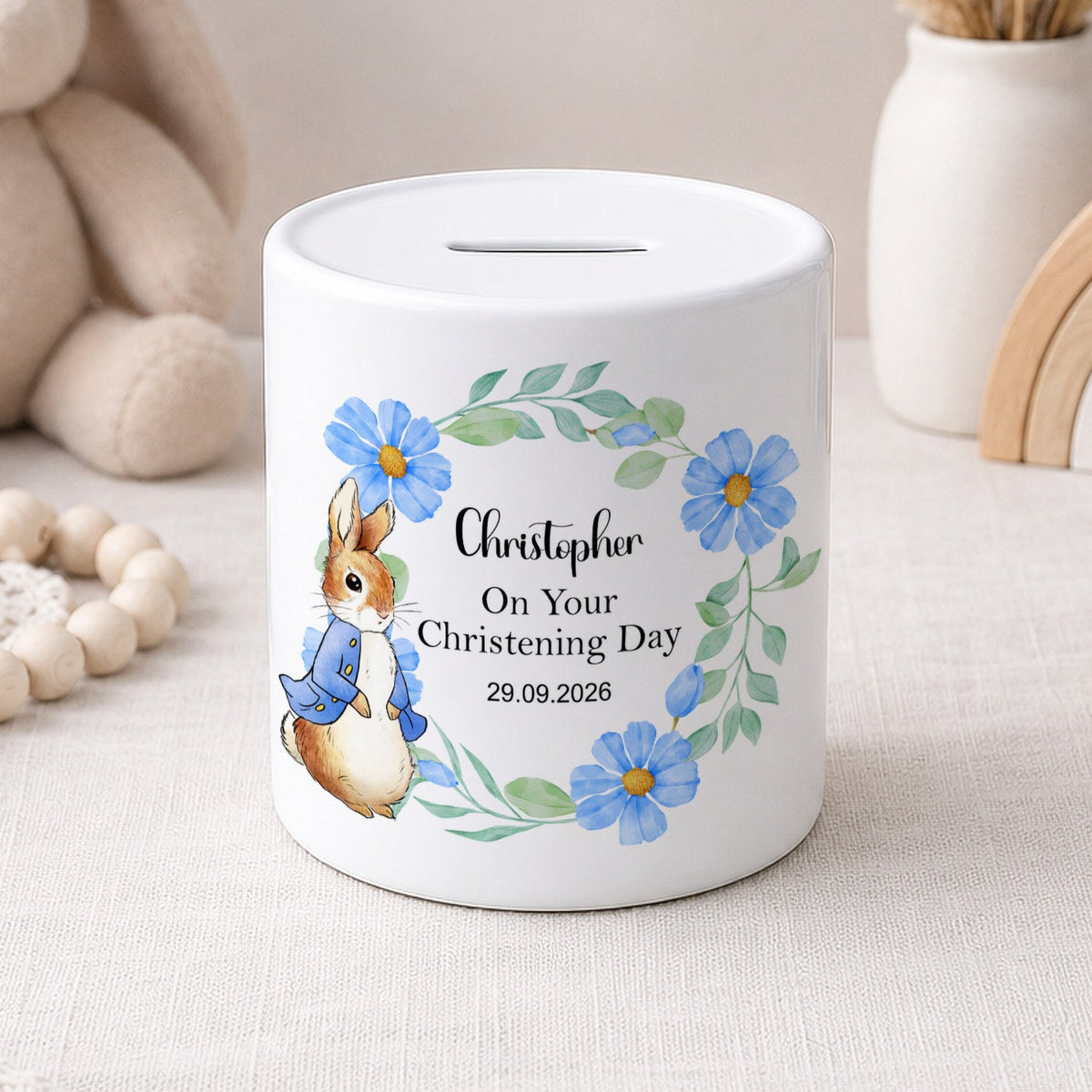 A white ceramic piggy bank with a personalised christening day design, featuring a watercolour rabbit in a blue outfit, blue flowers, and the name 'Christopher' on a light beige fabric surface with soft toys and decorative items in the background.