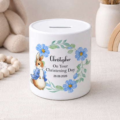 A white ceramic piggy bank with a personalised christening day design, featuring a watercolour rabbit in a blue outfit, blue flowers, and the name 'Christopher' on a light beige fabric surface with soft toys and decorative items in the background.