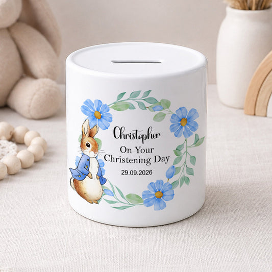A white ceramic piggy bank with a personalised christening day design, featuring a watercolour rabbit in a blue outfit, blue flowers, and the name 'Christopher' on a light beige fabric surface with soft toys and decorative items in the background.